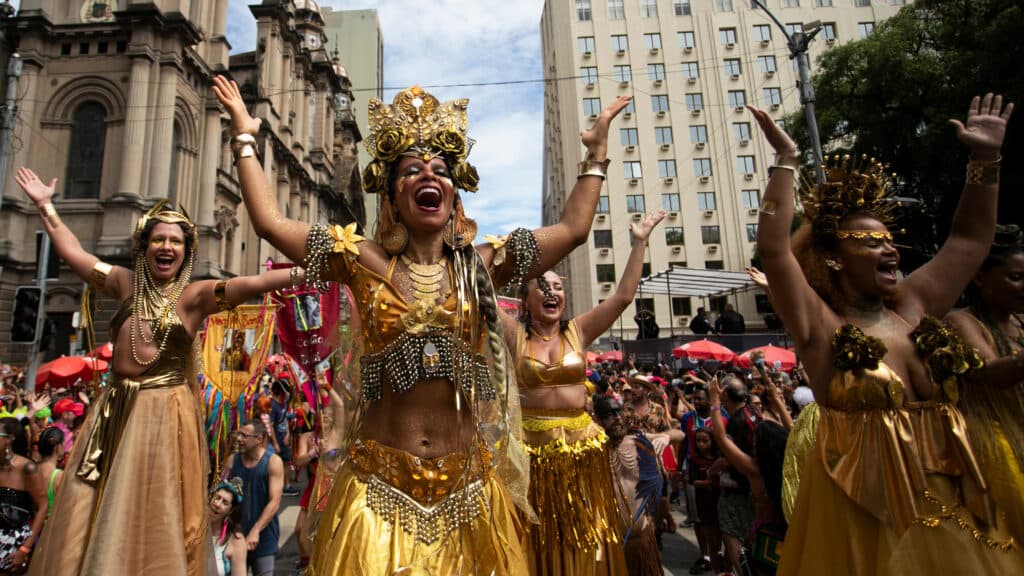 A group of people dressed in vibrant gold costumes with elaborate headdresses participate in a lively outdoor festival parade, with a historic building and a crowd in the background.