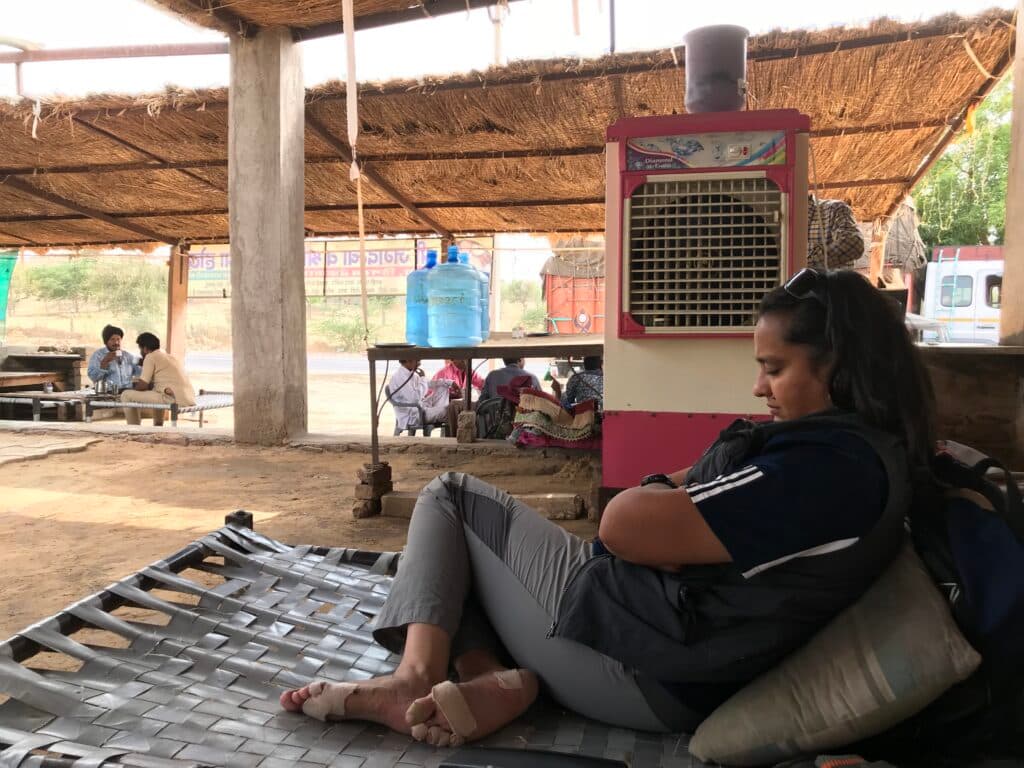 A woman resting on a woven cot under a canopy, with an air cooler and water containers nearby, while others are seated at tables in the background in an outdoor setting.