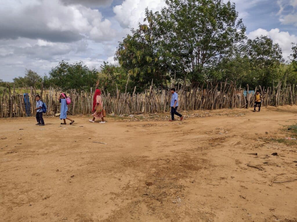 Children walking on a dirt path near a fence made of wooden sticks, with trees and a cloudy sky in the background.