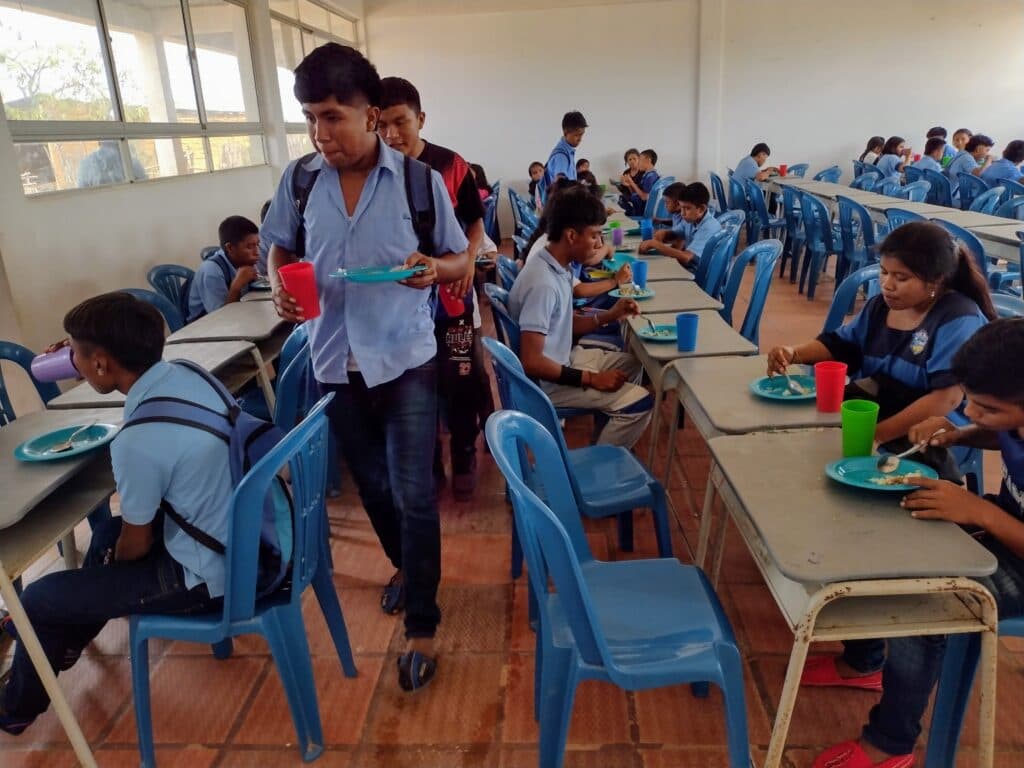 Students in uniforms eating lunch at long tables in a school cafeteria with blue chairs and light streaming in from large windows.