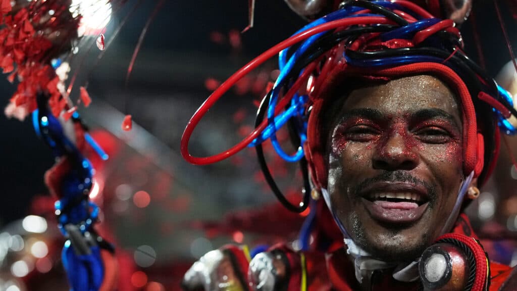 A person smiling, adorned with colorful red and blue decorations and glitter on their face, likely participating in a festive event or parade.