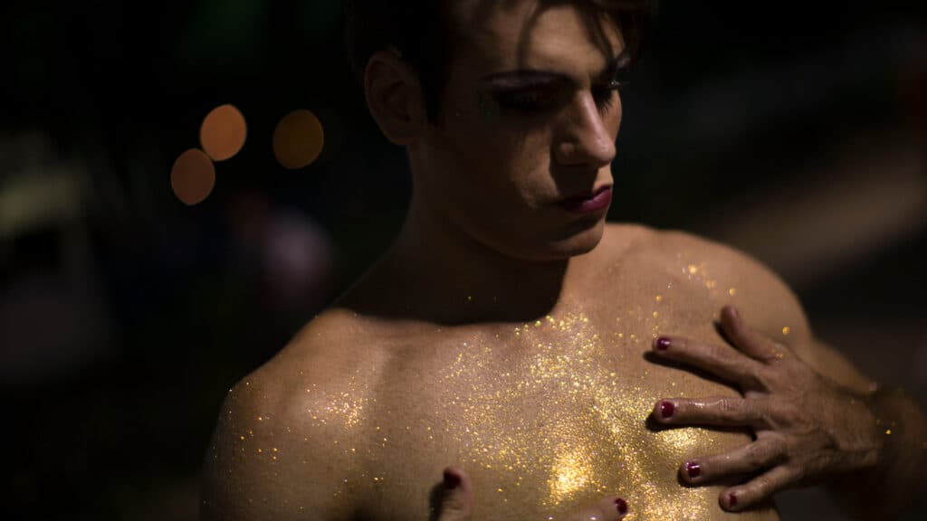 Close-up of a person with makeup and red-painted nails, covered in gold glitter on their upper body, against a dark background.