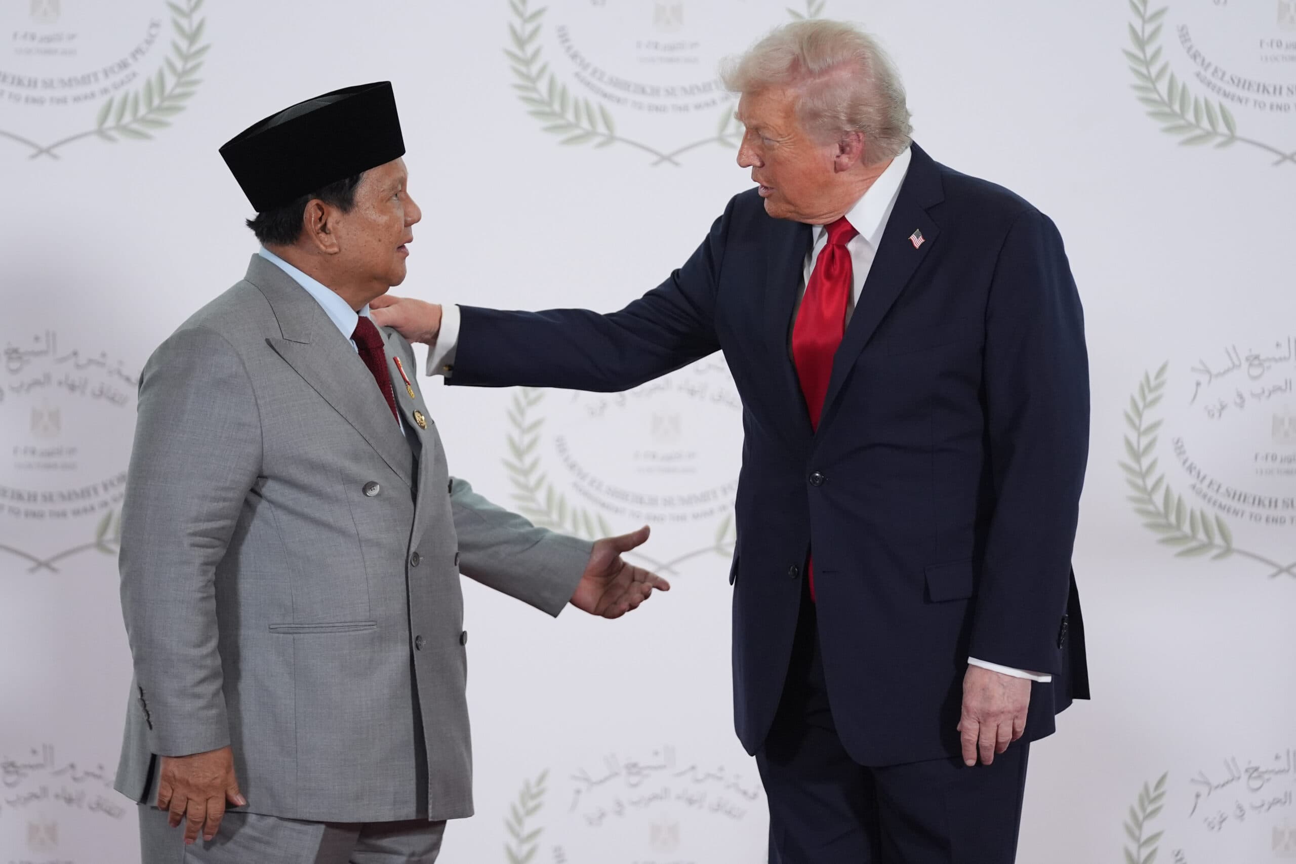Two men in formal attire, one wearing a gray suit and black cap, and the other in a navy suit with a red tie, are having a conversation in front of a backdrop with the words 'Sharm El-Sheikh Summit for Peace' and laurel wreaths. The man in the navy suit has his hand on the shoulder of the other man.