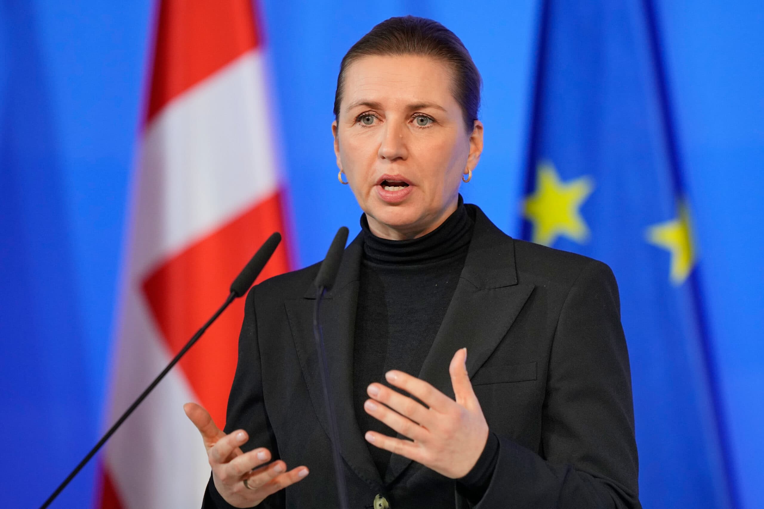 A woman in a black suit and turtleneck speaks into microphones with Danish and European Union flags in the background.