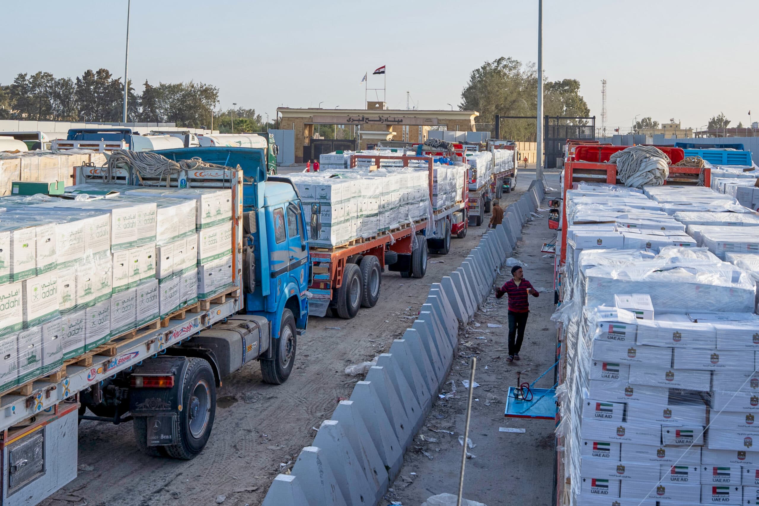 A row of trucks loaded with pallets of aid packages labeled "UAE Aid" parked along a barrier, with a child walking nearby, under a clear sky.