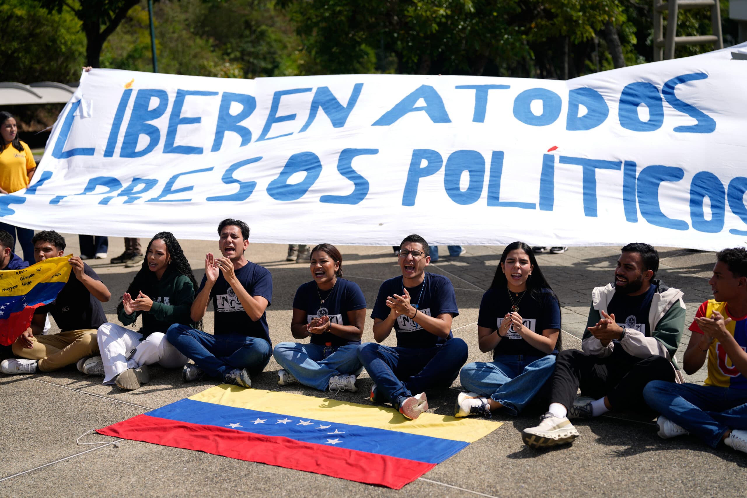 A group of people sitting on the ground, clapping, and holding a banner that reads 'Liberen a todos los presos políticos' in Spanish, with a Venezuelan flag displayed in front of them.