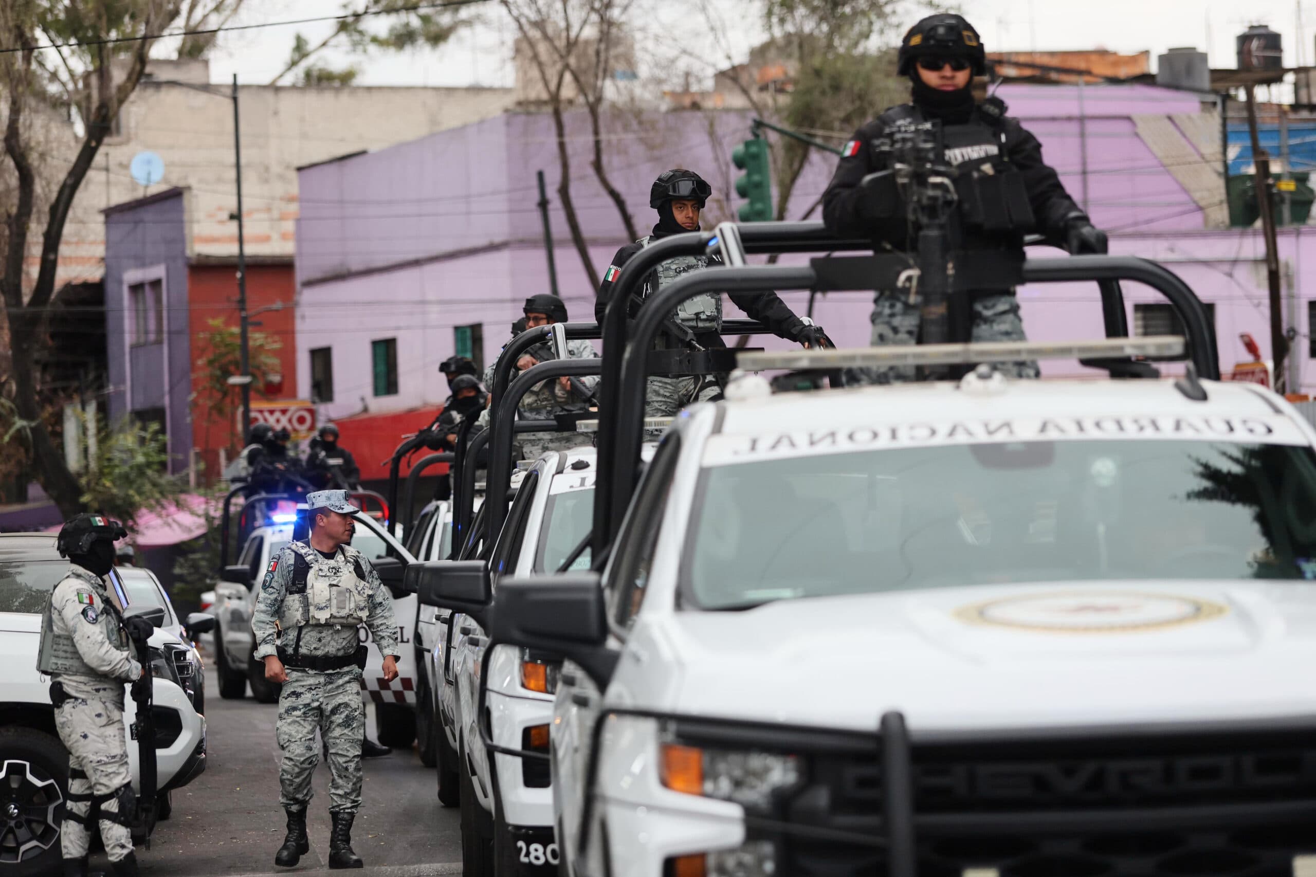 A group of uniformed security forces standing on and around several white patrol vehicles marked with "Guardia Nacional," in an urban setting with colorful buildings in the background.