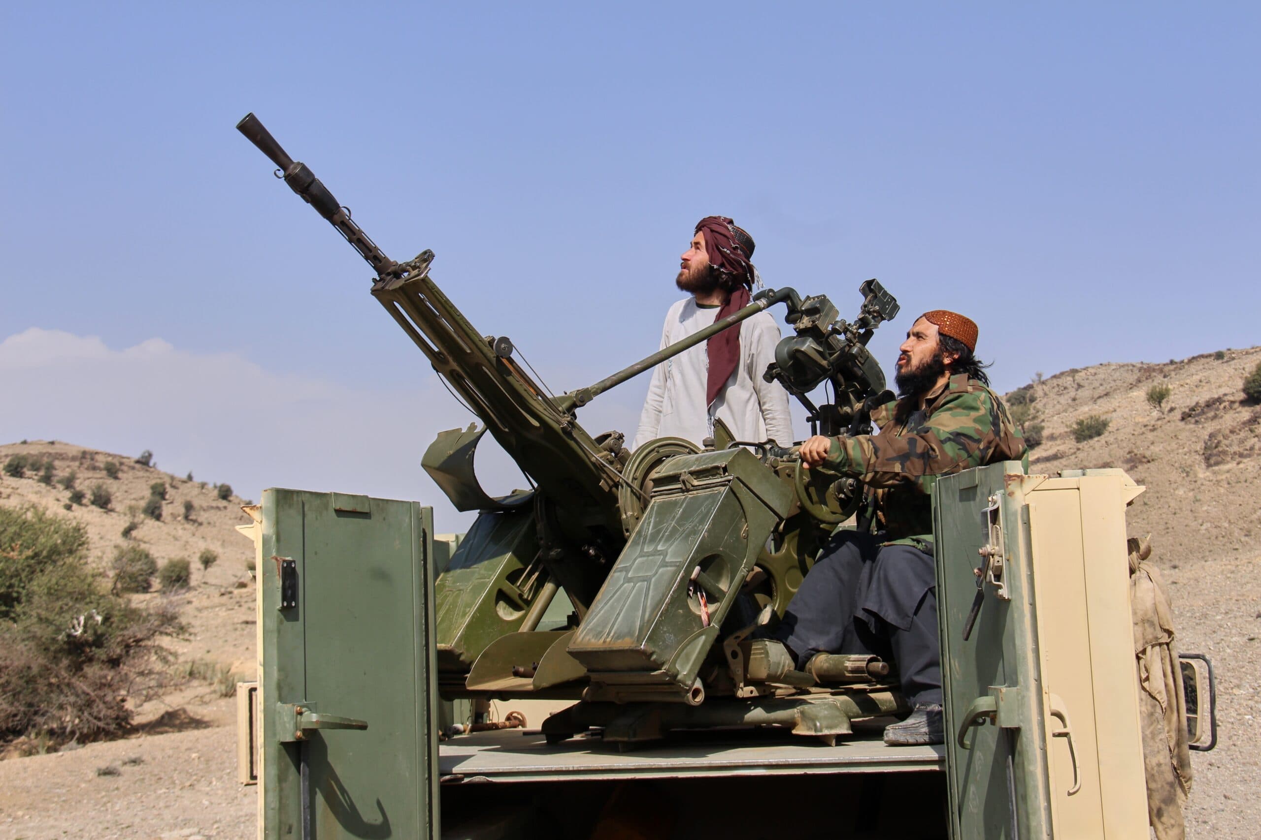 Two men in traditional and camouflage clothing operate an anti-aircraft gun mounted on a vehicle in a hilly, arid landscape under a clear blue sky.