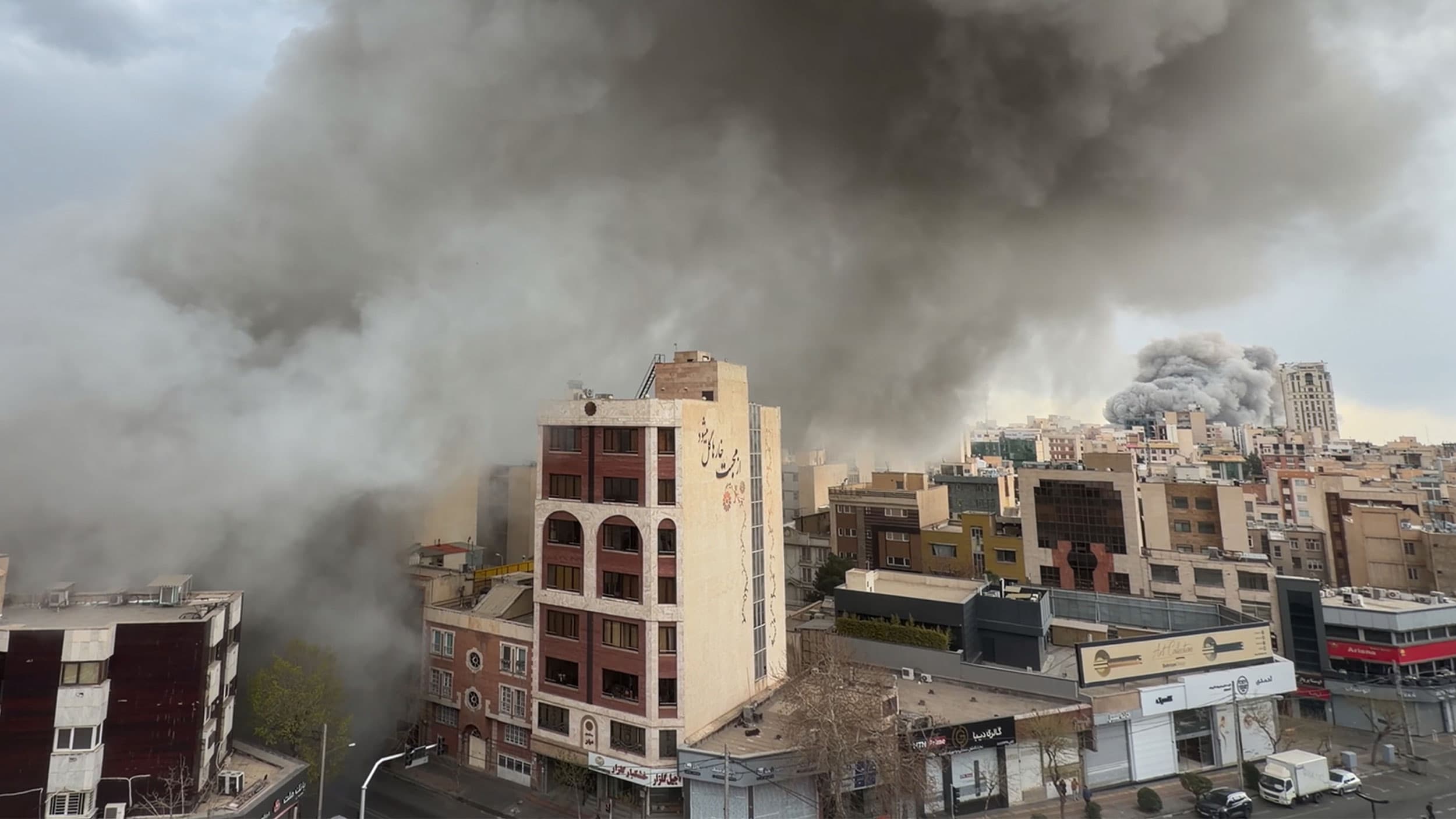 Large plumes of dark smoke rise above a cityscape, partially obscuring buildings against a gray sky, indicating a fire or explosion in an urban area.