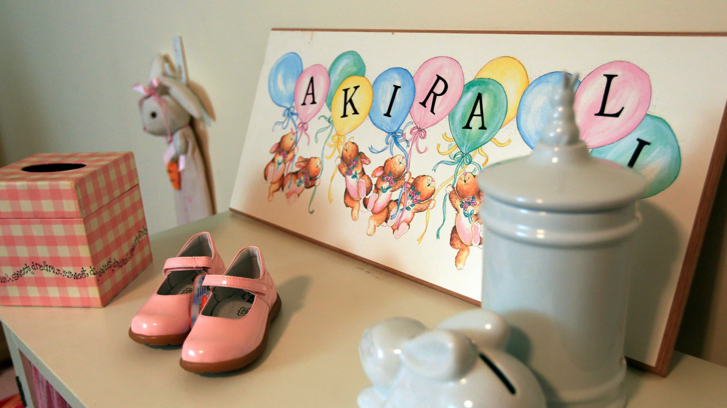 A child's room shelf with pink patent leather shoes, a pink plaid tissue box, and a framed illustration. The illustration depicts teddy bears holding balloons with letters forming a name. In the foreground, there is a pale blue container and a white ceramic item, possibly a toy or decoration. The setting is playful and colorful, suggesting a nursery or child's room.