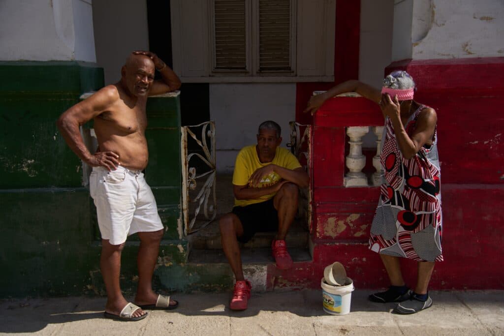 Three people in casual attire sit and stand outside a colorful building, with one person shirtless, another seated in a yellow shirt, and the third wearing a patterned dress and head scarf, covering their face.