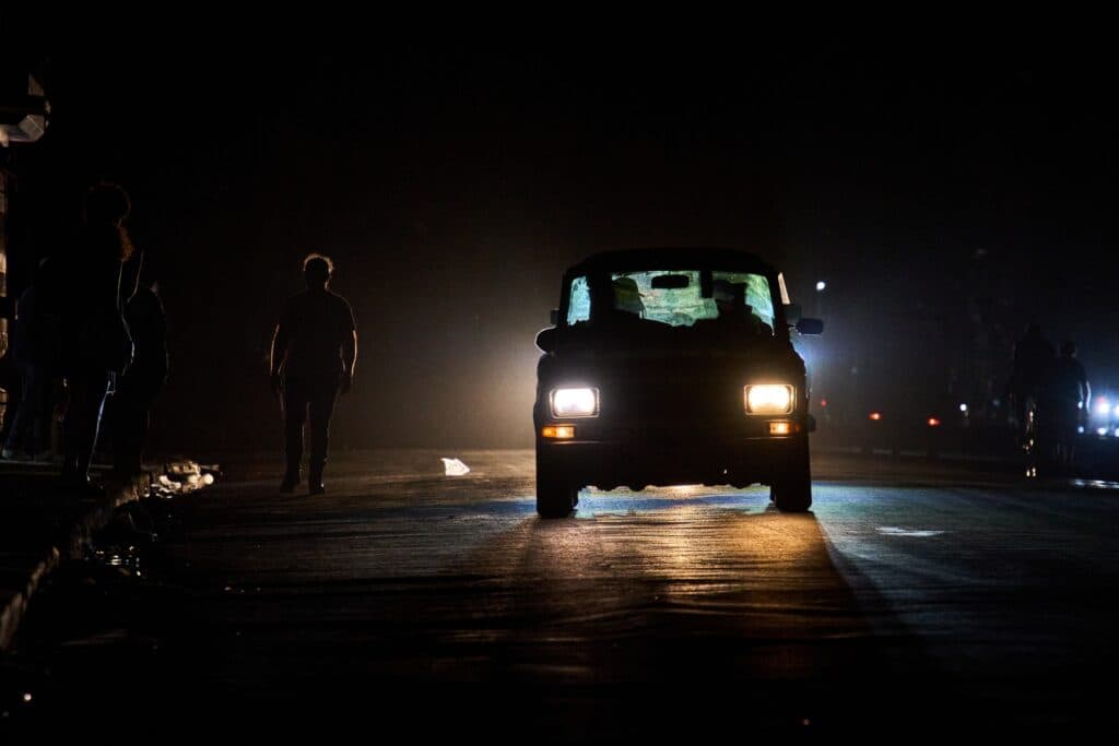 A nighttime scene with a car facing forward, headlights on, illuminating the street. A person walks on the road, silhouetted against the car's headlights. The background is dark, suggesting an urban environment.