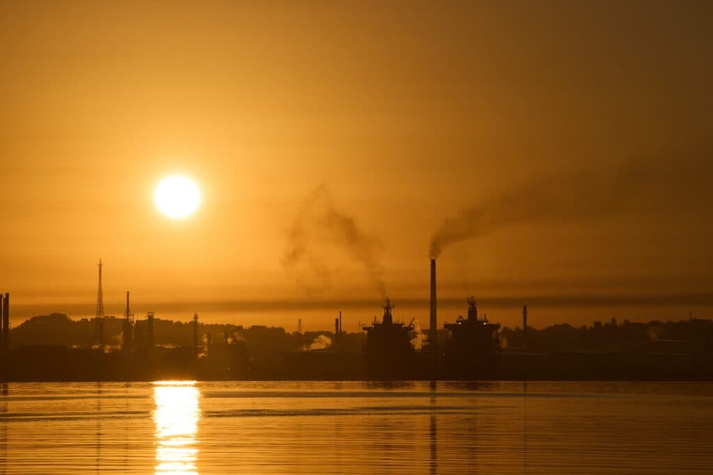 A sunset over an industrial area with smoke rising from chimneys, silhouetted against an orange sky with the sun reflecting on the water below.