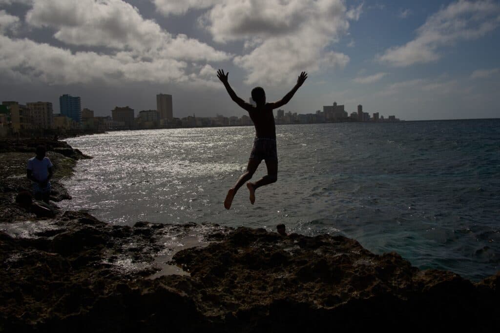 Silhouette of a person jumping off a rocky shoreline into the sea, with a city skyline in the background and sunlight reflecting on the water.