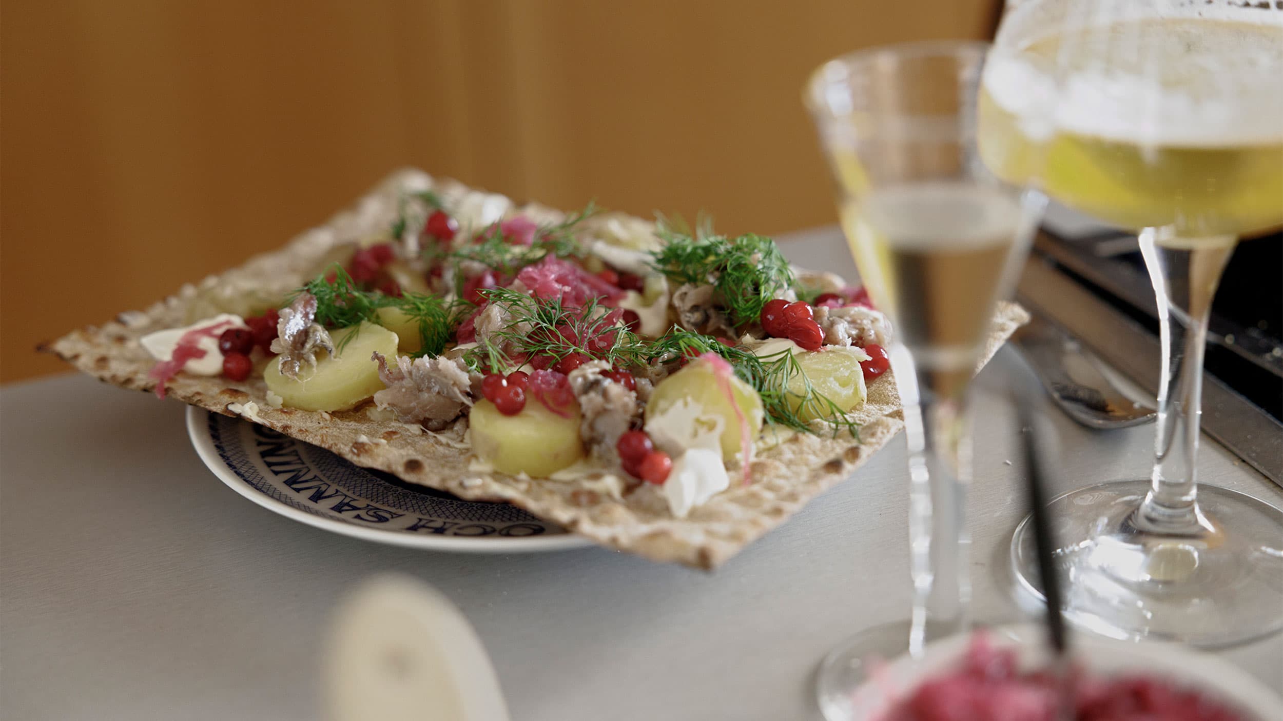 A plate of crispbread topped with potatoes, herring, red onions, dill, and red berries, accompanied by two glasses of beer.