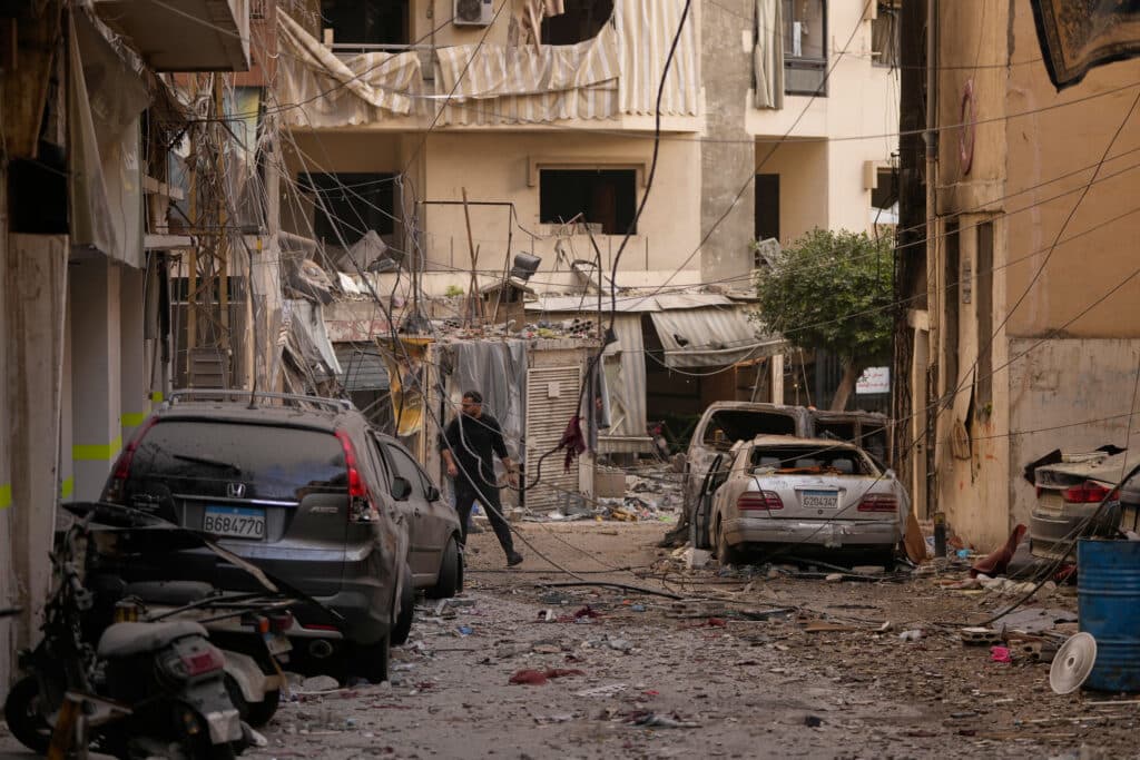 A street filled with debris, damaged vehicles, and rubble, showing the aftermath of destruction in an urban area. Buildings have broken windows and hanging awnings, with a person walking amidst the wreckage.