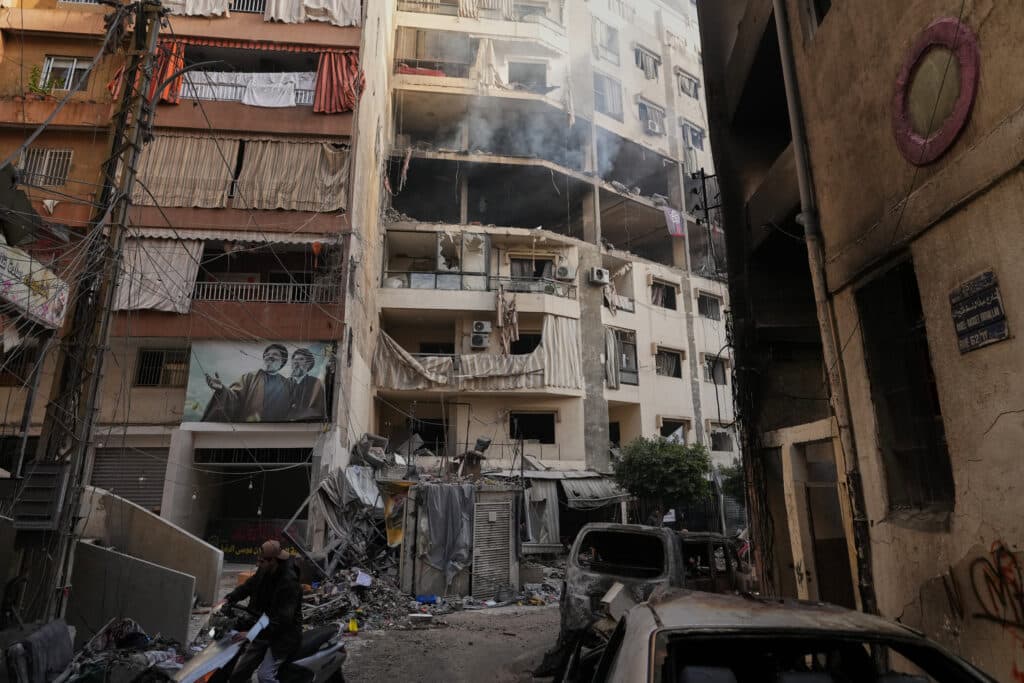 A damaged apartment building with smoke emanating from the upper floors, surrounded by debris and damaged vehicles, including a person walking past on a scooter. The building displays signs of destruction with broken windows, exposed interiors, and hanging cables.