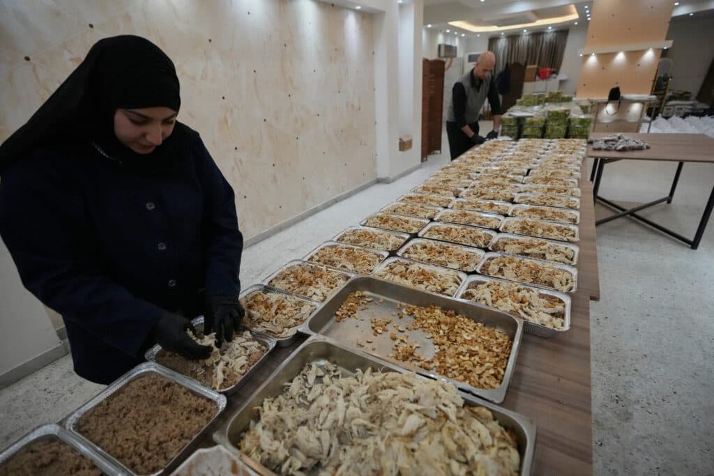 A woman and a man prepare and organize trays of food, including rice and chicken, on long tables in a large room; the trays are likely for a communal or charitable event.