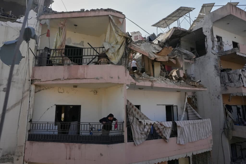 A damaged residential building with structural damage and debris, showing collapsed walls and ceilings. A person is visible on a balcony looking down, with tattered curtains hanging from the railing. Solar panels are visible on the rooftop.