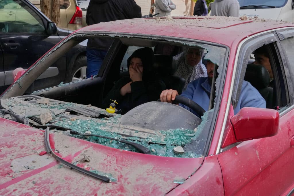 A red car with its windshield shattered and covered in debris, with multiple people inside; two women wearing headscarves in the back seat, and a man and a woman in the front seats, the latter covering her mouth with her hand.