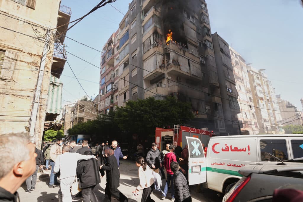 A multi-story residential building on fire with flames and black smoke emerging from a high floor. Below, people are gathered on the street, and emergency vehicles such as an ambulance and a fire truck are visible. The scene is busy with bystanders and responders amidst the backdrop of closely packed urban architecture.