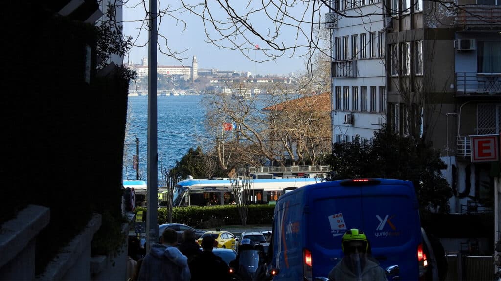 View down a narrow city street with a blue van and people in the foreground, leading to a waterfront with a tram and distant buildings across the water under a clear sky.