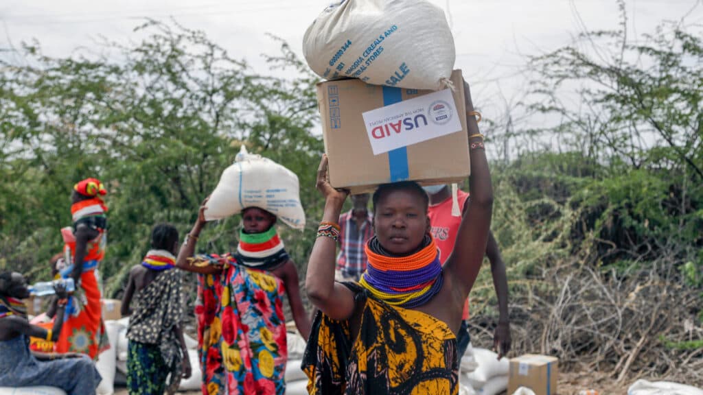 Women in colorful traditional clothing carry bags and a box labeled 'USAID' on their heads, surrounded by trees and bushes in a rural setting.