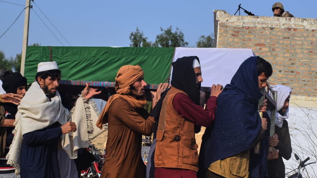 People carrying a green and white draped coffin on their shoulders during a funeral procession, with an armed guard on a rooftop in the background.