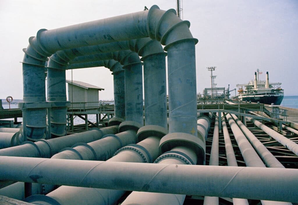A series of large oil pipelines run along a coastal structure, with a tanker ship docked in the background.