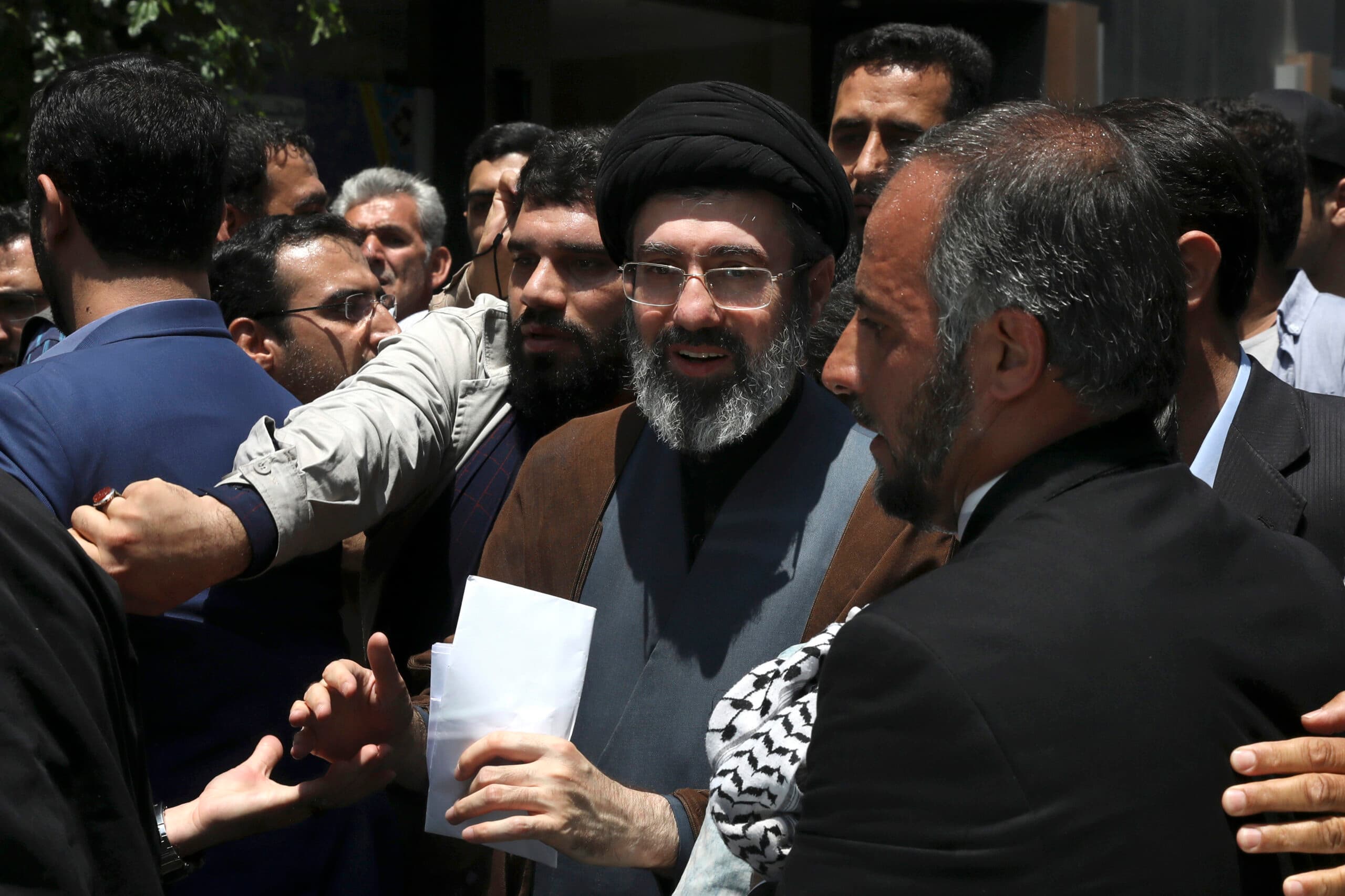 A group of men, including a central figure wearing a black turban and glasses, are gathered closely together, with some extending their arms in gestures. The man in the turban holds a white paper.