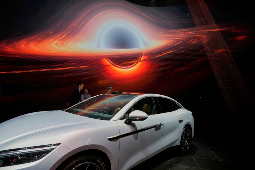 A sleek white car is displayed indoors against a backdrop of a vivid cosmic scene featuring a bright, swirling black hole displayed on a large screen. Several people are visible in the background, observing the display.
