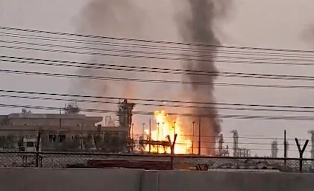A large fire with flames and smoke rising from an industrial facility, viewed from a distance with power lines and fences in the foreground.