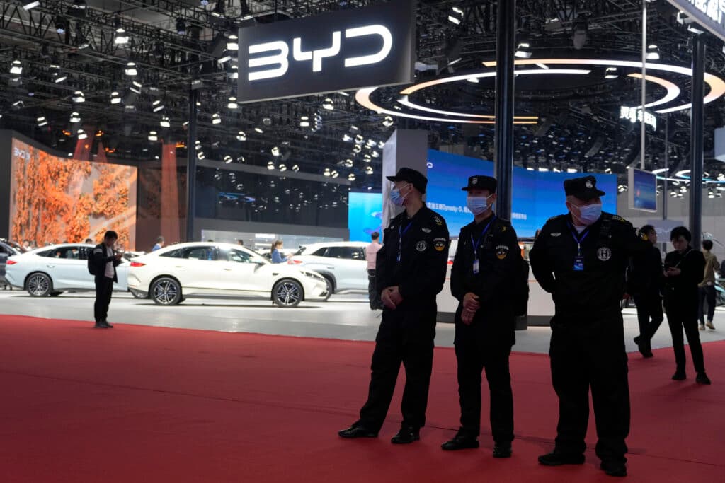 Several people stand in a car exhibition area with BYD signage, featuring white cars on display and uniformed individuals wearing face masks in the foreground.