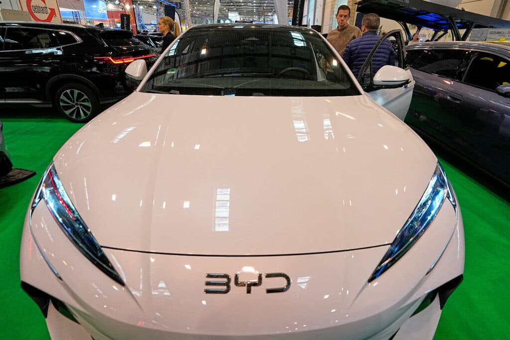 Front view of a white BYD electric car displayed at an auto show on a green carpet, surrounded by other vehicles and attendees.