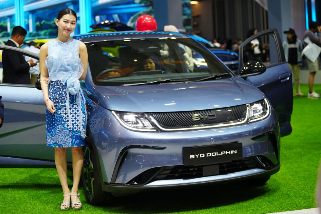 A person stands next to a blue BYD Dolphin car displayed at an event, with people in the background. The car is parked on artificial grass, and the setting appears to be a car show or exhibition.