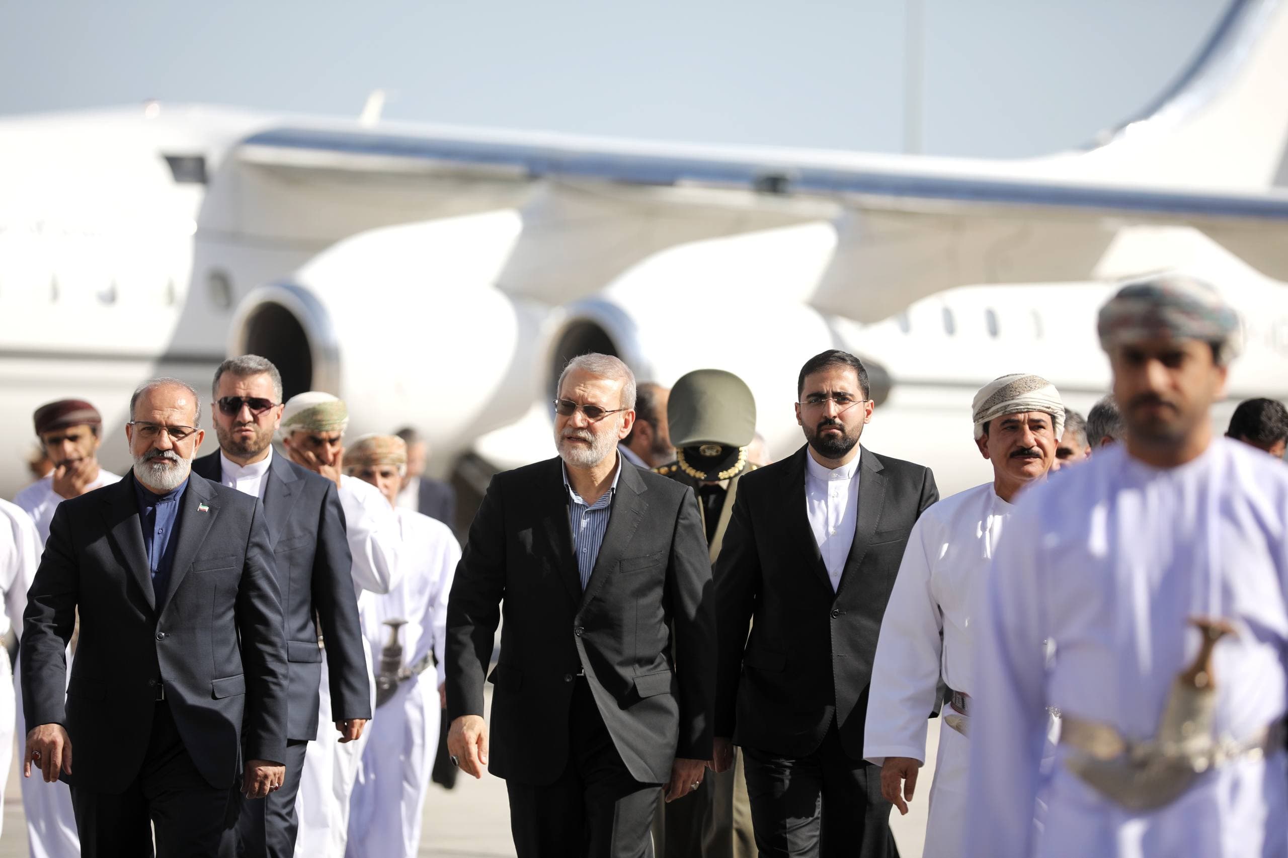 A group of men in formal attire walk on an airport tarmac with a plane in the background.