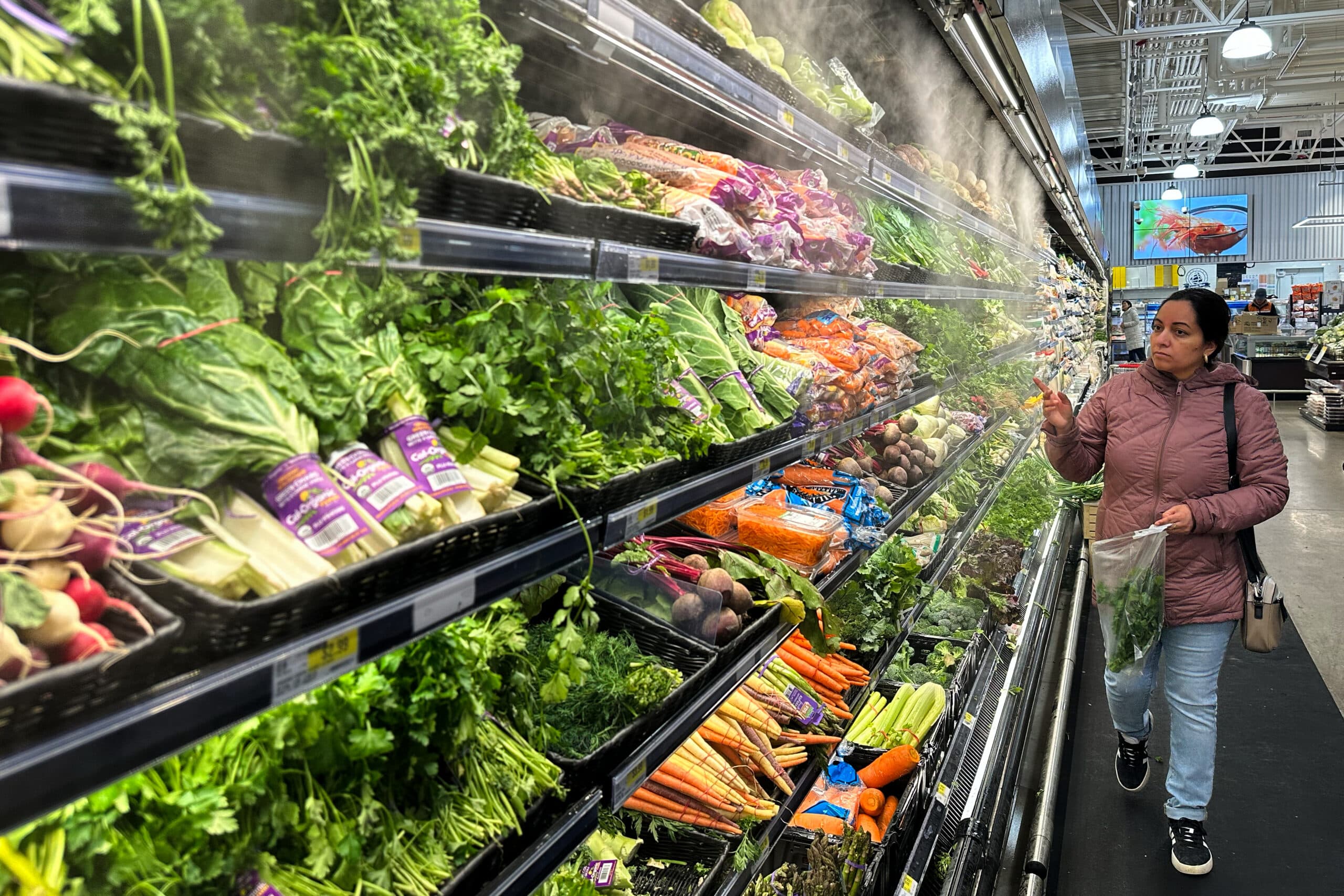 A woman shopping in a grocery store, selecting fresh produce from a misted display case filled with various vegetables like celery, carrots, and radishes. She is wearing a pink jacket and holding a bag with greens in it.