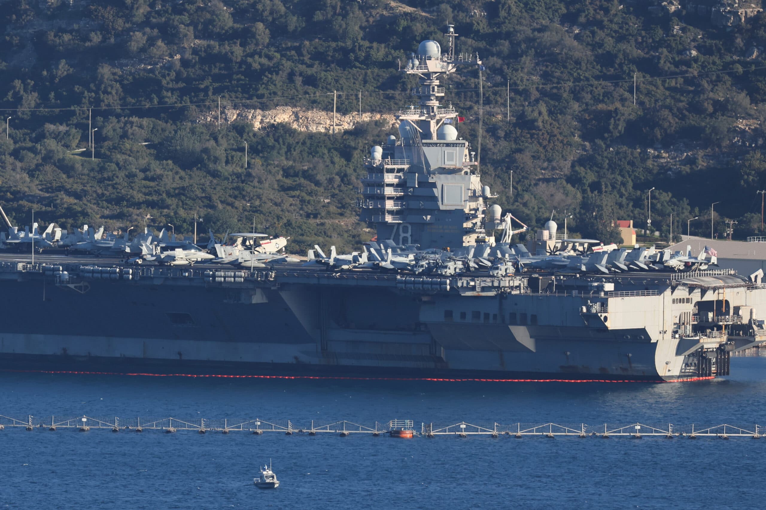 A large aircraft carrier stationed in a body of water, with multiple fighter jets visible on its deck, set against a backdrop of wooded hills.