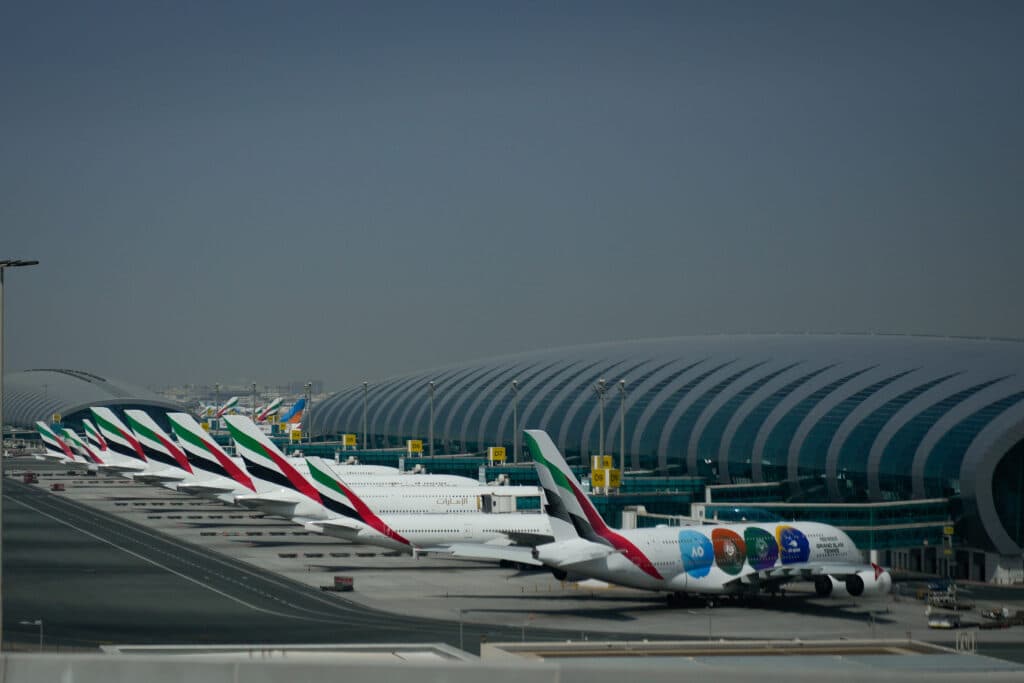 Several airplanes with Emirates livery are parked at a modern airport terminal with a distinctive arched roof.