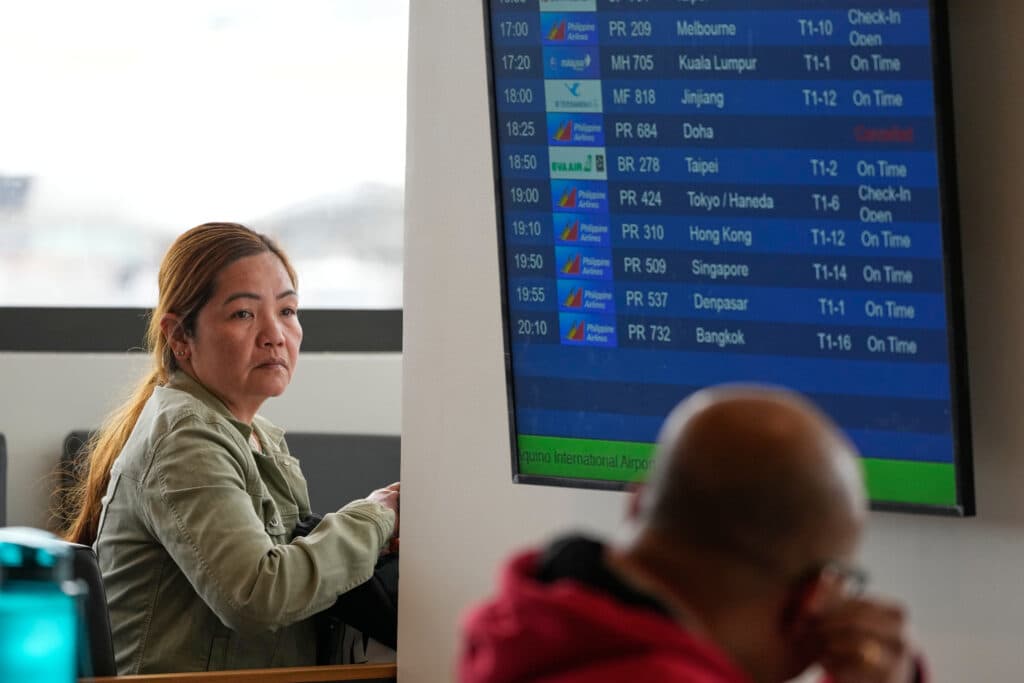 A woman looking at a flight information display board at an airport, showing various flights and their statuses, including one canceled flight to Doha.