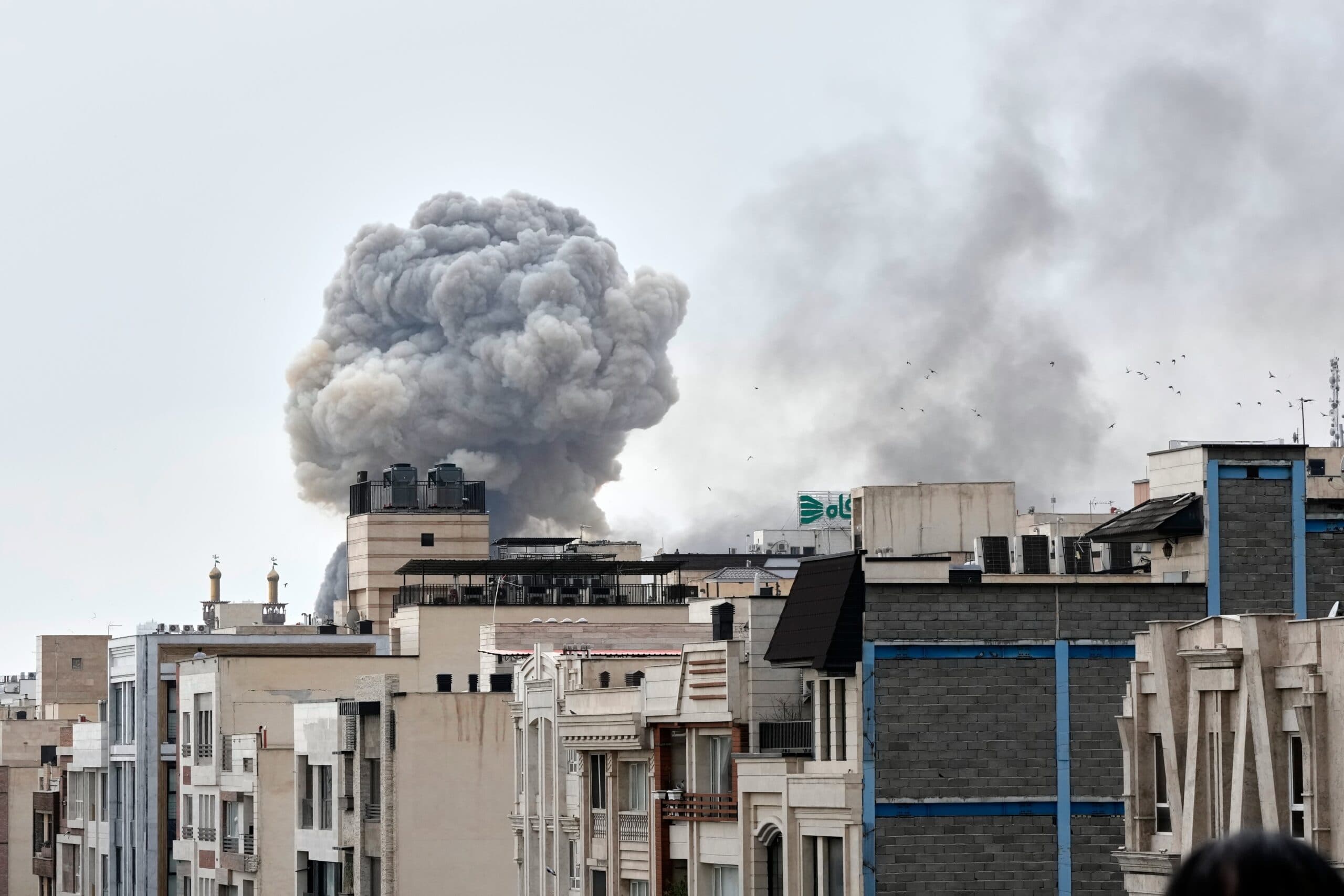 Large plume of smoke rising from behind buildings in a city landscape.