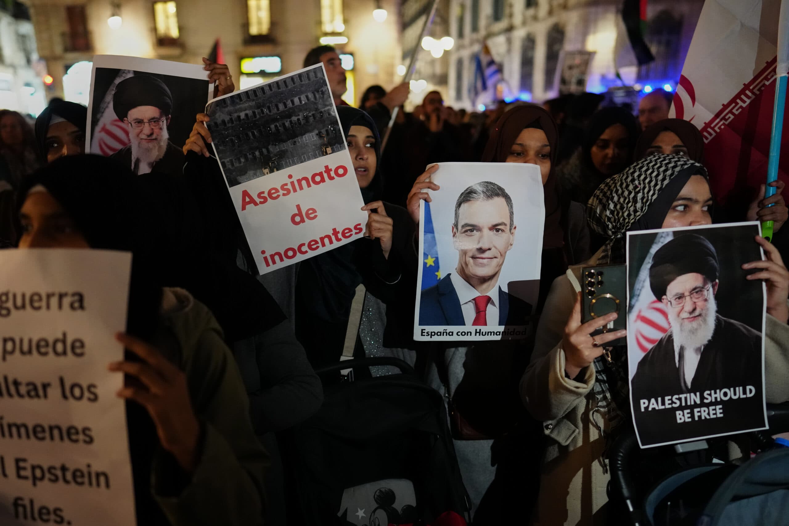A group of people holding protest signs, including images and messages such as "Asesinato de inocentes" and "Palestine should be free," in a nighttime setting with buildings in the background.