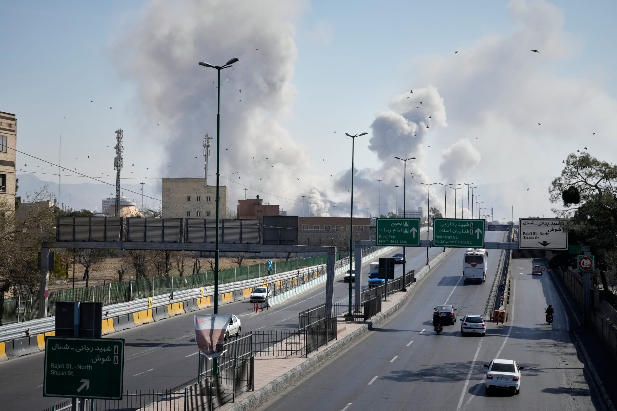A cityscape view of a wide road with few vehicles and large plumes of smoke rising in the background, suggesting an explosion or fire. Directional signs in both Arabic and English are visible, and there are some birds flying in the blue sky.