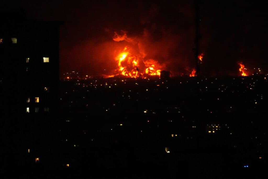 A cityscape at night with large fires burning, creating a bright orange glow against the dark sky. A silhouetted building with illuminated windows is visible in the foreground.