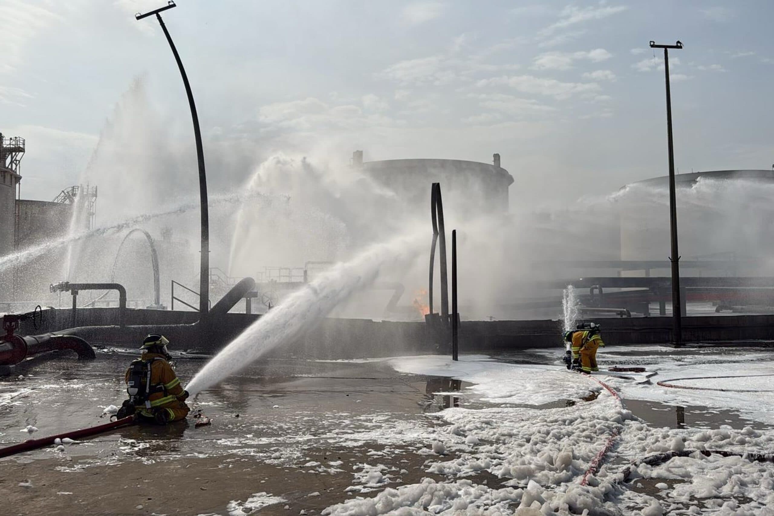 Firefighters spray water and foam onto a fire at an industrial facility surrounded by large storage tanks, with hoses on the ground and mist in the air.