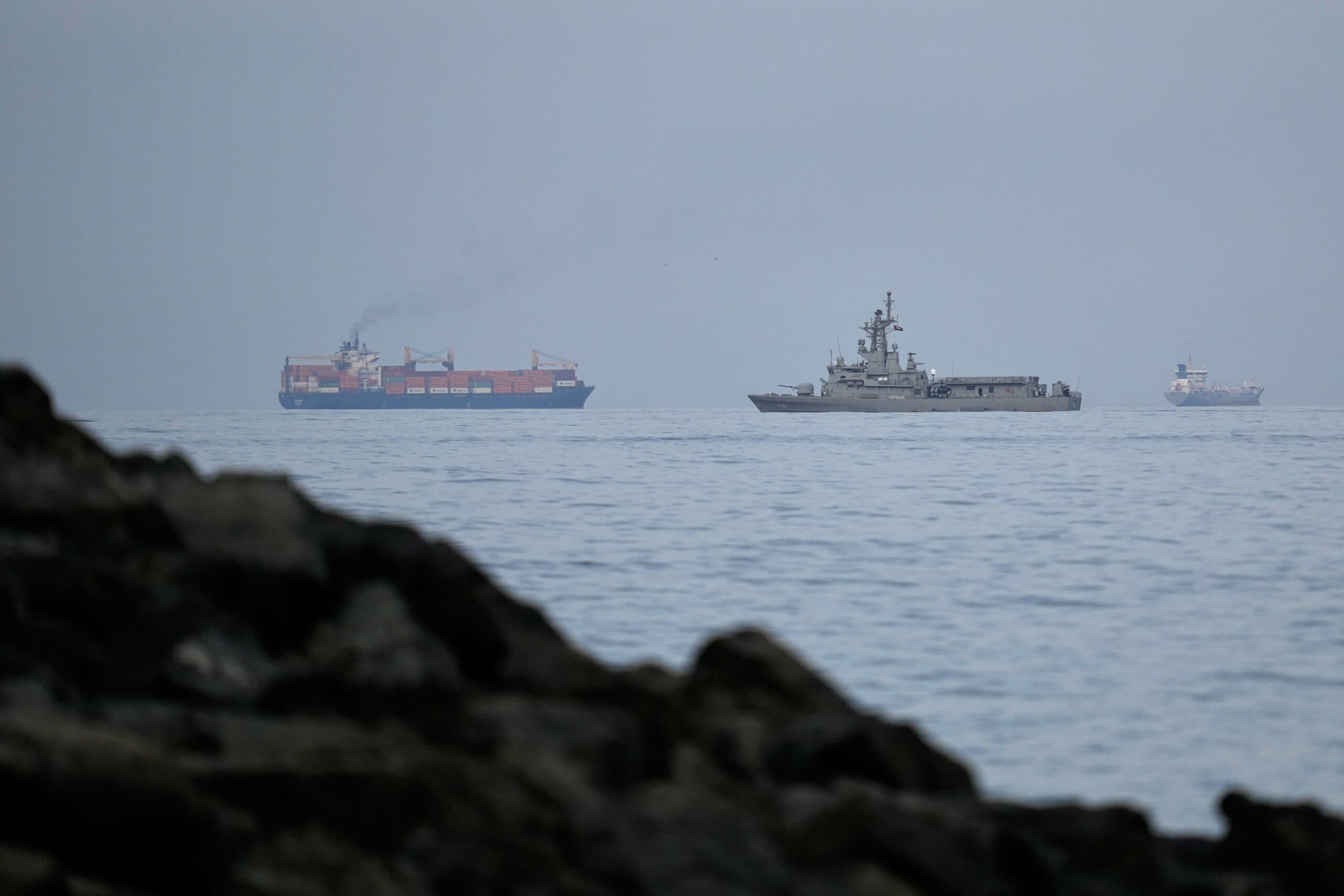 A naval ship sailing near two cargo ships in calm waters, with a rocky shoreline in the foreground.