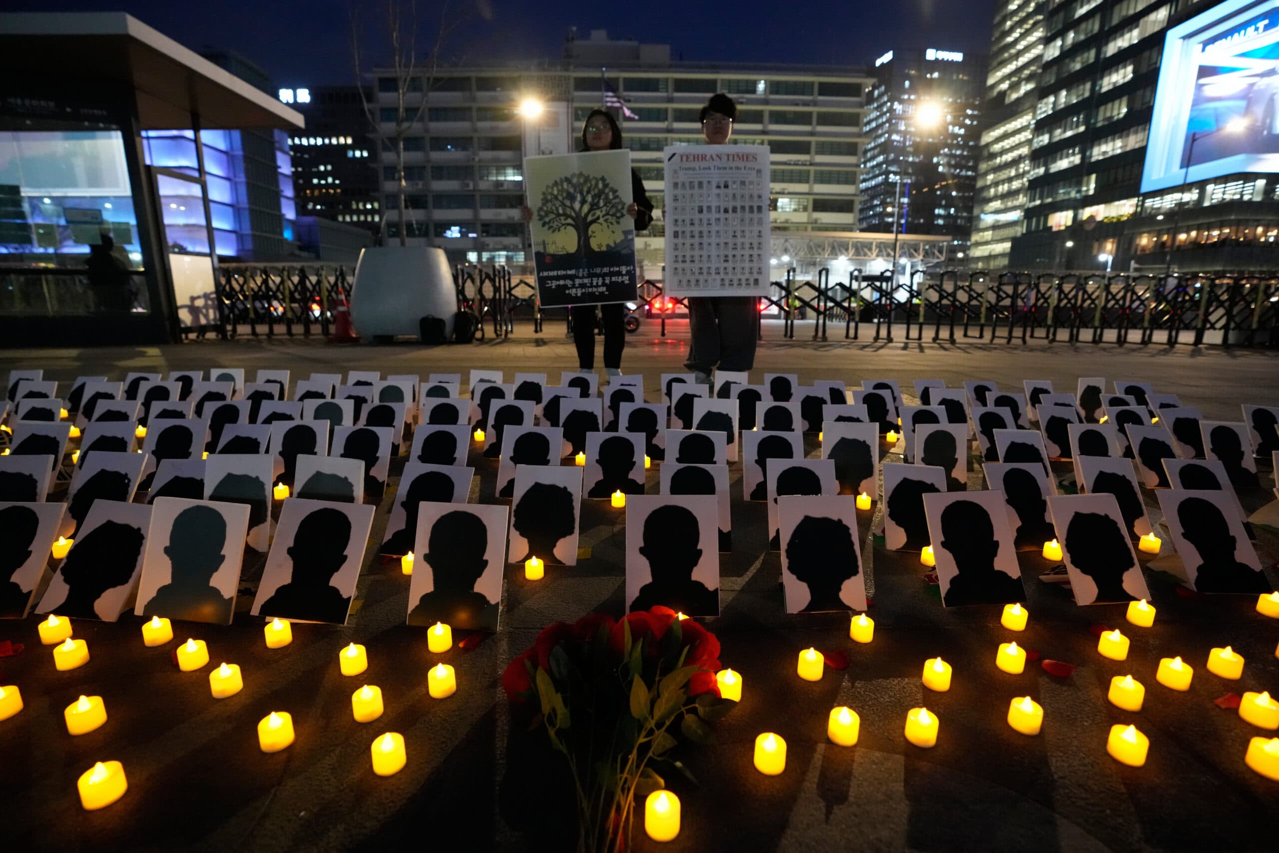 An outdoor vigil at night features numerous silhouette portraits placed on the ground, surrounded by glowing candles. Two people stand holding posters, with a backdrop of city buildings and ambient evening lighting.