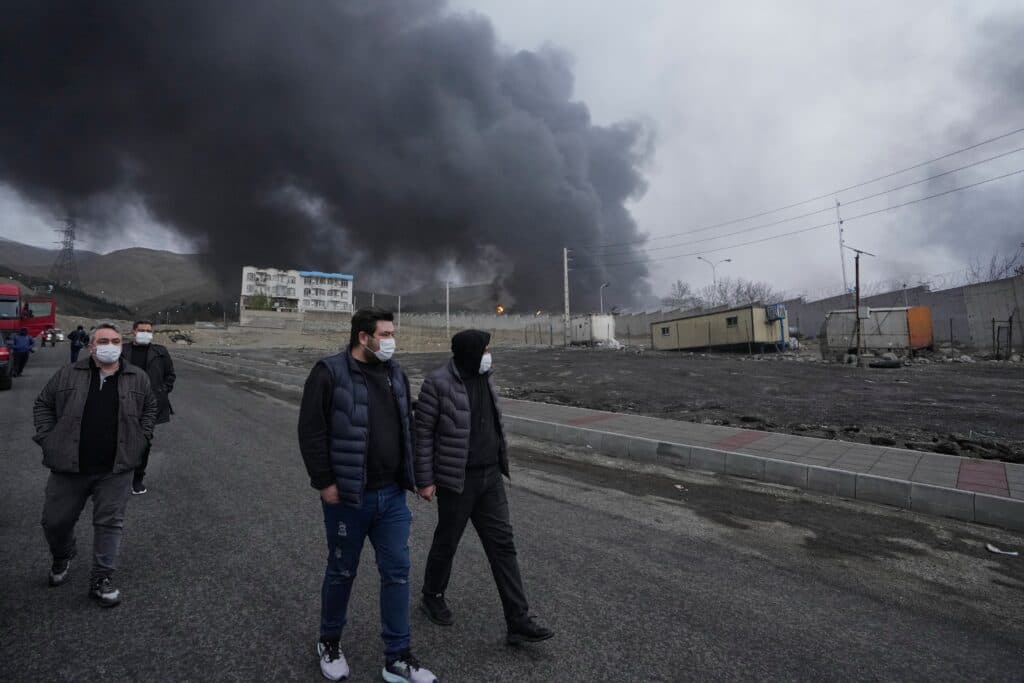 People wearing masks walk on a road with a large plume of black smoke in the background, indicating a fire near industrial buildings and hills.