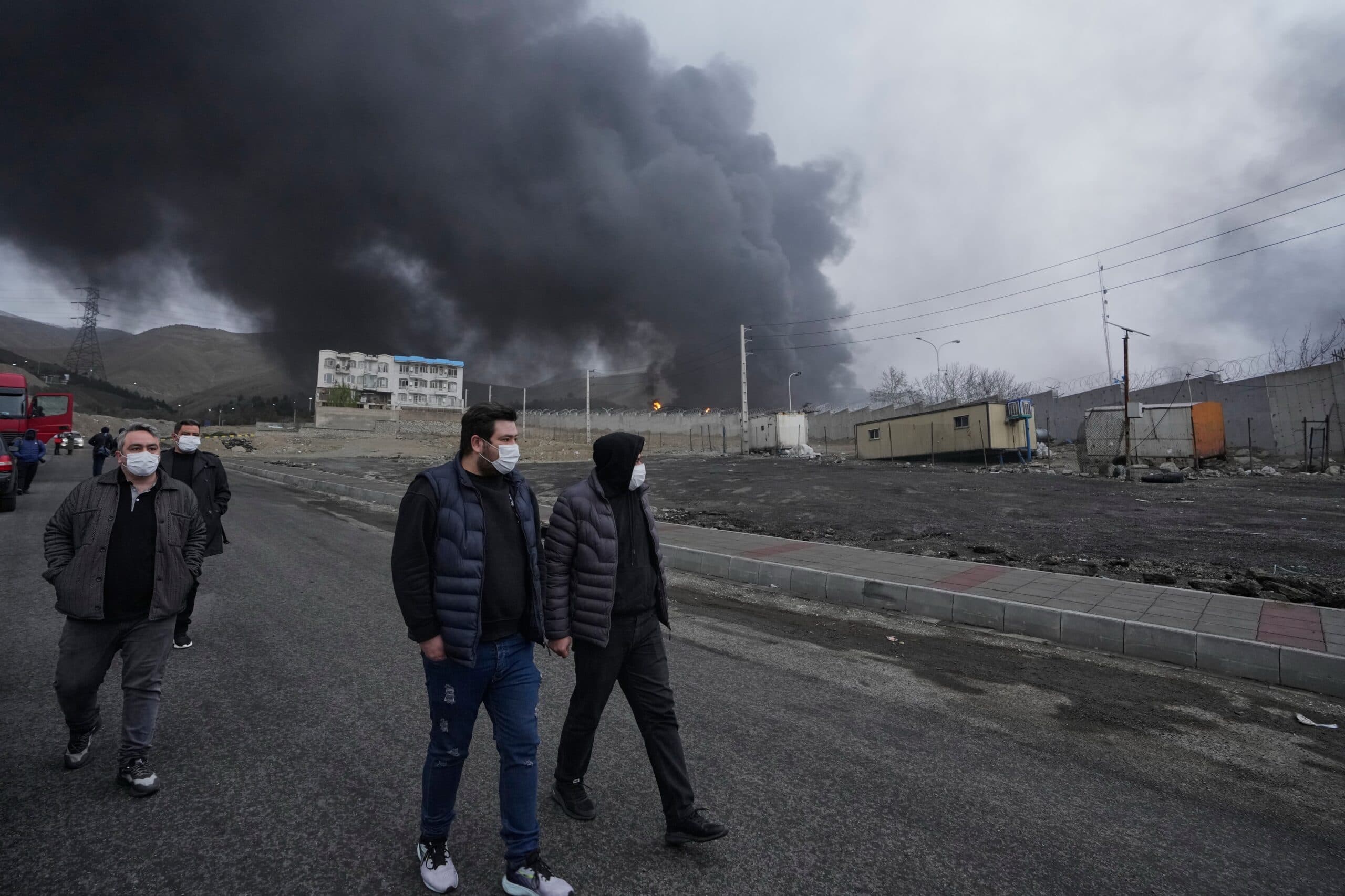 People wearing masks walking along a road with thick black smoke rising in the background from an industrial area, with containers and a building visible nearby.