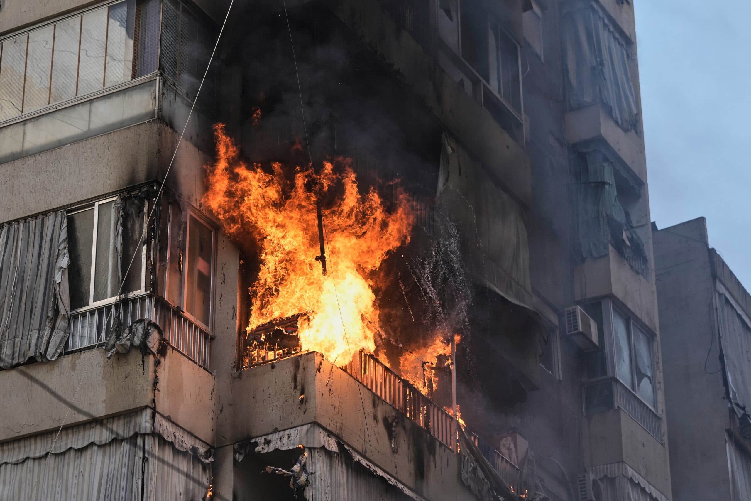 A large fire engulfing a balcony of a multi-story apartment building, with flames and smoke rising from the structure.